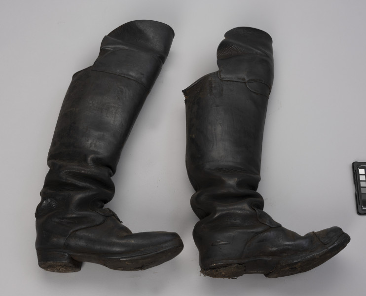 tłok pieczętny - Ujęcie z boku; Para butów rybackich o wysokich cholewach zszytych z dwóch kawałków skóry, które od przodu sięgają powyżej kolan (ochraniacze). Drewniane podeszwy są podbite grubą, barwioną na kolor czarny, sztywną, prawdopodobnie impregnowaną tłuszczem, skórą za pomocą gwoździków. Czubek prawego buta jest wzmocniony kawałeczkiem skóry, a lewy ma wzmocnione skórą palce.