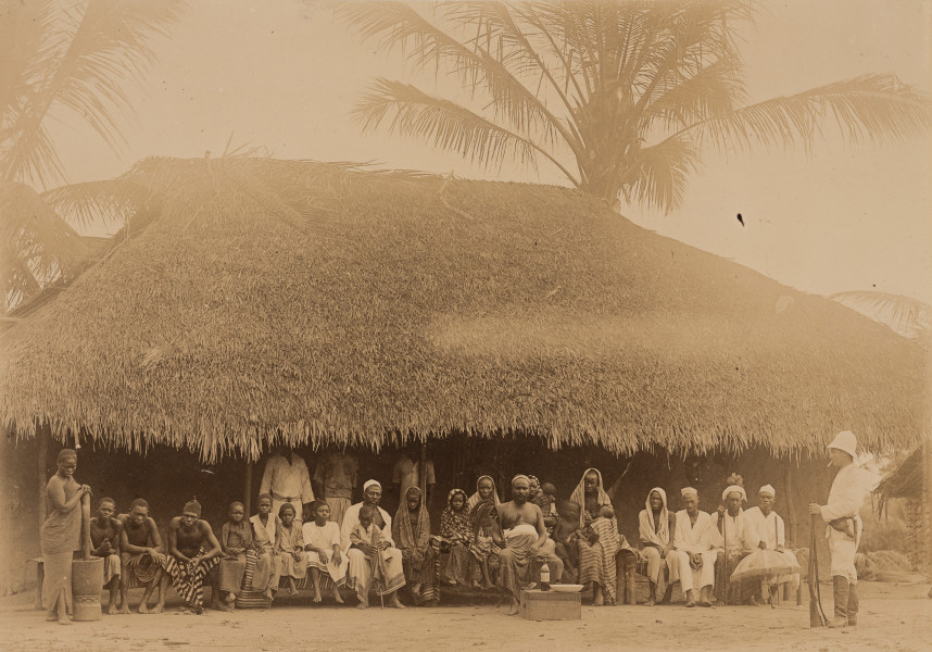 fotografia z albumu Aleksandra Dandelskiego; König Ringu mit Gefolge (König Kingu mit Gefolge?) - ujęcie z przodu; Fotografia w układzie poziomym przedstawia miejscowego króla wraz ze świtą. Król o endomorficznej budowie ciała, siedzi na tronie lekko wysuniętym do przodu, przed grono osób mu towarzyszących, w dłoni trzyma insygnia władzy. Za nim na połączonych ławkach posadzono jego świtę: miejscowe kobiety, mężczyzn i dzieci. Bezpośrednio za nim znajdują się trzy kobiety wraz z dziećmi, ubrane w charakterystyczne spódnice z chustami zakrywającymi głowy – być może są to żony króla. Po obu stronach kobiet posadzono mężczyzn ubranych w białe stroje i nakrycia głowy, trzymających w dłoniach laski lub parasole. Najprawdopodobniej są to miejscowi dostojnicy, poważana starszyzna. Następnie siedzą dzieci i sądząc po ubiorze mężczyźni o niższej randze, być może rzemieślnicy. Obok stoi miejscowa kobieta. Trzyma w ręku stępor, który włożony jest do stępy. Prawdopodobnie przerwano jej pracę podczas rozdrabniania ziaren. Za siedzącą świtą stoi trzech ubranych na biało miejscowych mężczyzn, ich twarze zasłania pokryty strzechą dach stanowiącej tło zabudowy. Obok siedzących dostojników, po prawej stronie, stoi ubrany w biały mundur i hełm oficer, w dłoni trzyma broń palną, kolbą opiera ją o ziemię, a w ustach ma papierosa. Przed królem ustawiono obróconą do góry dnem skrzynię, na której postawiono szklaną butelkę oraz biały głęboki talerz. Ponad zabudową widać również liście palm.