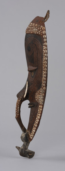 maska mai - Ujęcie z prawej; Drewniana maska antropomorficzna z długim nosem ozdobionym kłami dzikiej świni oraz gliniano-drewnianym mostkiem zwanym molot. Maska wydłużona i szczupła, stosunkowo wąska twarz z długim nosem, szerokimi ustami. Pod wydatnymi łukami brwiowymi znajdują się skośne oczy (w tym ujeciu widoczne tylko jedno) z zaznaczonymi powiekami, o kącikach skierowanych ku górze i gałkach ocznych w postaci muszli. Molot ma formę łukowatego uchwytu zaczynającego się poniżej nozdrzy, pod ozdobą nosa, a sięgającego do brody zakończonej ciosaną głową ptaka. Maska pokryta brązowymi barwnikami, wpadającymi w rdzawy i ciemnobrązowy kolor. Zdobiona wzorami geometrycznymi w postaci prostych i zakręcających linii (jest to prawdopodobnie forma tatuażu). Całość mai otoczona została czerwoną gliną w której umieszczono muszle, m.in. nossa i kauri (porcelanki). W ten sposób zaznaczono również policzki, opaskę nad czołem oraz górną część mostka. Na szczycie umieszczono pleciony uchwyt.