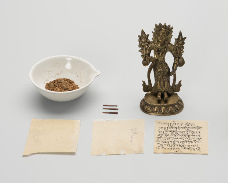 figurka Bodhisattwa Awalokiteśwara Padmapani wraz z wypełnieniem - ujęcie z przodu; Z prawej strony mała, wykonana z brązu figurka przedstawiająca Bodhisattwę Awalokiteśwarę Padmapani wraz z wypełnieniem w postaci trzech zwitek papieru, rozłożone na płasko z przodu (dwie zapisane, jedna czysta), z lewej strony figury trzy patyczki (patyczek w trzech częściach? - najprawdopodobniej był to jeden patyczek na który zawinięto arkusze papieru), obok w miseczce materiał sypki na który składa się: zawartość roślinna (ryż, nasiona) oraz glina - przechowywane w osobnej kopercie, kartoniku i woreczku strunowym. Postać ludzka o skupionej, lekko uśmiechniętej twarzy, na głowie diadem. Stoi na lotosowym postumencie o 11 parach odwróconych płatków, którego górną część ozdobiono paciorkami. Ubrana jest w szaty królewskie: przewiązaną w talii tkaninę z paskiem od którego odchodzą łańcuszki z paciorków oraz biżuterię książęcą: trójramienny diadem, parę, dużych, okrągłych kolczyków, podwójny naszyjnik z paciorków z wisiorkami, bransolety na nadgarstkach i przedramionach. Do prawego ramienia przylega kwiat lotosu (typowy atrybut), natomiast po lewej stronie, symetrycznie, na tej samej wysokości, przylega przypominający kwiat lotosu niezidentyfikowany atrybut Awalokiteśwary. Obie dłonie ułożone są w geście argumentacji: kciuk i palec wskazujący złączone, tworzą krąg. Prawa uniesiona jest na wysokości klatki piersiowej, lewa opada wzdłuż ciała, zatrzymując się na wysokości biodra. Z ramion figurki zwisa przewieszona skóra antylopy.