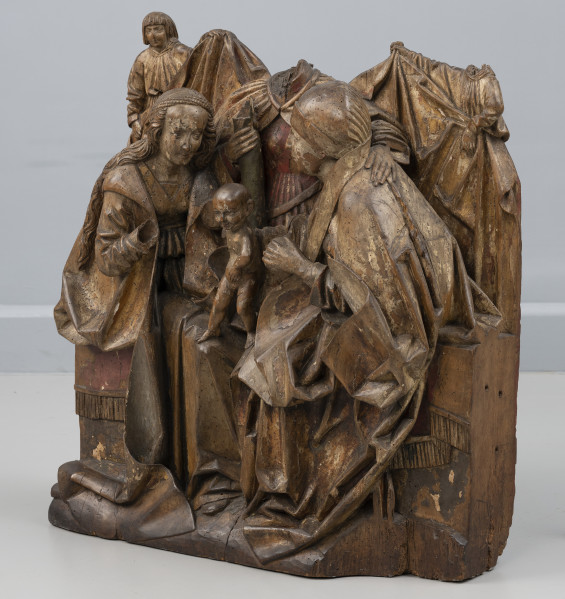 grupa rzeźbiarska - Ujęcie ze skosu z prawej; Grupa rzeźbiarska o zwartej, symetrycznej kompozycji, wykonana z jednego bloku drewna i polichromowana. Przedstawia trzy siedzące kobiety zwrócone ku dzieciątku stojącemu w centrum na ich kolanach. Analogicznie upozowane w układzie antytetycznym sylwetki długowłosej Marii po lewej stronie i Anny w czepcu po prawej okrywają obszerne płaszcze, układające się wokół figur dużymi płaszczyznami i opadające na podstawę sceny. Na ich kolanach stoi nagie dzieciątko o gładko opracowanej główce. Za nim, widoczna pomiędzy kobietami, siedzi frontalnie Emerancja o nieco powiększonych proporcjach, w prawej dłoni trzymając fragment pnia. Tło stanowi kotara unoszona przez dwa anioły widoczne w półpostaci, ubrane w szerokie tuniki. Tron okrywa tkanina obszyta frędzlami. Cechy anatomiczne postaci i modelunek ciężkich obszernych tkanin oddane są naturalistycznie. Rzeźba posiada znaczące ubytki – brakuje głów Emerancji i prawego anioła, dłoni Marii i prawej rączki dzieciątka oraz fragmentu podstawy z prawej strony. Na powierzchni rzeźby widoczne otwory po owadach. Polichromia zachowana częściowo.