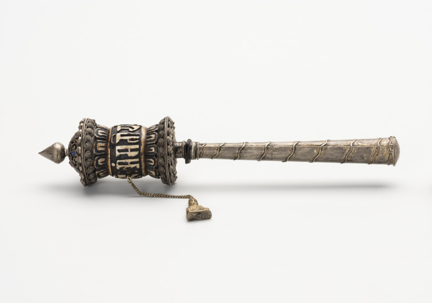 młynek modlitewny wraz z tekstem modlitewnym - Ujęcie z boku z lewej; Klepsydrowy, ręczny młynek modlitewny z uchwytem w formie tulejki w kształcie ściętego, wydłużonego stożka składający się z się z pięciu elementów oraz arkusz z tekstem modlitewnym niewidocznym na prezentowanym ujęciu. Mosiężny pąk lotosu u szczytu, kościano-mosiężna, klepsydrowa część z łańcuszkiem i inskrypcją oraz mosiężna podkładka tworzą bęben młynka. Poniżej znajdują się trzy kolejne elementy: niewielka, kościana podkładka, mosiężny uchwyt, z którego wystaje ostry, stalowy szpikulec. Młynek ozdobiono sześcioma, kamieniami pokrytymi niebieską emalią na zdjęciu widoczne tylko trzy z sześciu oraz motywami ślimacznicy, roślinnym, stylizowanych przecinków, zaokrąglonych linii i okręgów, zlokalizowanymi na mosiężnej powłoce bębna. Wymienione wzory wykonano z niklowanej blaszki mosiężnej, pociętej na drobne paseczki, przypominają zdobienie granulacją. Na kościanej części bębna wyrzeźbiono dwa razy słowa mantry: 