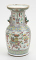 wazon typu famille rose - ujęcie z tyłu; Porcelanowy wazon zdobiony wielobarwną dekoracją naszkliwną w pastelowych odcieniach z przewagą różu w typie famille rose. Kształt gruszkowaty, szyja szeroka zakończona rozszerzającym się wylewem, w którym widoczny jest w tyle półokrągły ubytek. Wazon bogato zdobiony licznymi motywami dekoracyjnymi. W jego górnej części po bokach znajdują się dwa uchwyty w formie stylizowanych lwów chińskich w kolorze pastelowej zieleni. Brzeg wylewu zdobiony dookolnie i motywem stylizowanego lotosu o czerwonym kwiecie, żółtych i zielonych liściach na białym tle oraz zgeometryzowanym wzorem roślinnym w kolorach zielonym i różowym na żółtym tle. Szyję oddzielono od brzuśca biegnącymi dookolnie wzorami: ornamentem meandrycznym o różowej, żółtej i seledynowej barwie oraz znajdującym się pod nim motywem grzyba nieśmiertelności w niebieskim, żółtym, czerwonym i seledynowym kolorze. Brzusiec oraz szyję zdobią czworoboczne pola (rezerwy) o seledynowych bordiurach wypełnione tzw. motywem Stu Antyków (Hundred Antiques/Hundred Treasures) na białym tle. Ułożono je na na szyi oraz na brzuścu. Pola wypełniają przedmioty i rośliny w dość nieformalnym układzie. Na szyi wyodrębnić można: różnorodne wazy (z przykrywkami oraz bez, na stojaku oraz bez), dyski z jadeitu, podwójną tykwę, z której unosi się smużka dymu oraz inne niezidentyfikowane przedmioty. Na brzuścu pokazano: wazę z kwiatami na stojaku, akwarium na stojaku, różnorodne wazy, donicę z miniaturową rośliną na stojaku, naczynie na wysokim stojaku oraz inne niezidentyfikowane przedmioty i rośliny. Dół wazonu ozdobiony dookolnie geometrycznym motywem w postaci pionowych żółtych i różowych pasków na seledynowym tle. Na brzuścu widoczne są ślady klejenia.