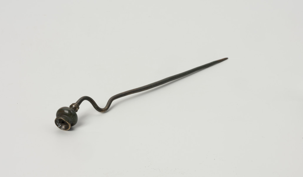 szpila z brązu ze Świchowa - Ujęcie z przodu; Szpila z brązu z wygiętą esowato  łabędzią szyjką. Zwieńczenie stanowi główka w formie miniaturowej wazy, której podstawę stanowią 3 dookolne zgrubienia w formie pierścieni. Powierzchnia zabytku jest w bardzo dobrym stanie, pokrywa ją ciemnozielona patyna.