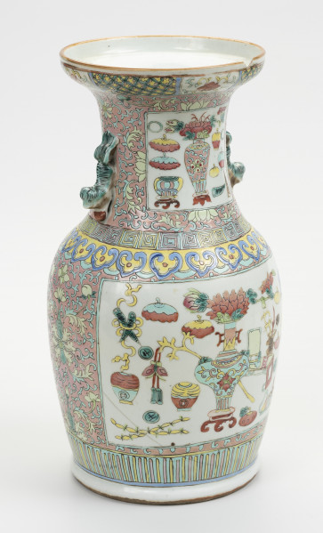 wazon typu famille rose - ujęcie z przodu z lewej; Porcelanowy wazon zdobiony wielobarwną dekoracją naszkliwną w pastelowych odcieniach z przewagą różu w typie famille rose. Kształt gruszkowaty, szyja szeroka zakończona rozszerzającym się wylewem, w którym widoczny jest z prawej półokrągły ubytek. Wazon bogato zdobiony licznymi motywami dekoracyjnymi. W jego górnej części znajdują się dwa uchwyty w formie stylizowanych lwów chińskich w kolorze pastelowej zieleni. Brzeg wylewu zdobiony dookolnie i naprzemiennie motywami stylizowanego lotosu o czerwonym kwiecie, żółtych i zielonych liściach na białym tle oraz zgeometryzowanym wzorem roślinnym w kolorach zielonym i różowym na żółtym tle. Szyję oddzielono od brzuśca biegnącymi dookolnie wzorami: ornamentem meandrycznym o różowej, żółtej i seledynowej barwie oraz znajdującym się pod nim motywem grzyba nieśmiertelności w niebieskim, żółtym, czerwonym i seledynowym kolorze. Brzusiec oraz szyję zdobią czworoboczne pola (rezerwy) o seledynowych bordiurach wypełnione tzw. motywem Stu Antyków (Hundred Antiques/Hundred Treasures) na białym tle. Pola wypełniają przedmioty i rośliny w dość nieformalnym układzie. Na szyi wyodrębnić można: wazon z peoniami na stojaku, różnorodne wazy (z przykrywkami oraz bez, na stojaku oraz bez), akwarium na stojaku, oraz inne niezidentyfikowane przedmioty. Na brzuścu pokazano:  wazę z kwiatami na stojaku, akwarium na stojaku, parę skrzyżowanych rogów nosorożca, toaletkę z lustrem, różnorodne wazy, donicę z miniaturową rośliną na stojaku, naczynie na wysokim stojaku oraz inne niezidentyfikowane przedmioty i rośliny. Tło stanowi przedstawiony na różowym tle motyw stylizowanego lotosu z wijącymi się liśćmi. Dół wazonu ozdobiony dookolnie geometrycznym motywem w postaci pionowych żółtych i różowych pasków na seledynowym tle. Na brzuścu widoczne są ślady klejenia.