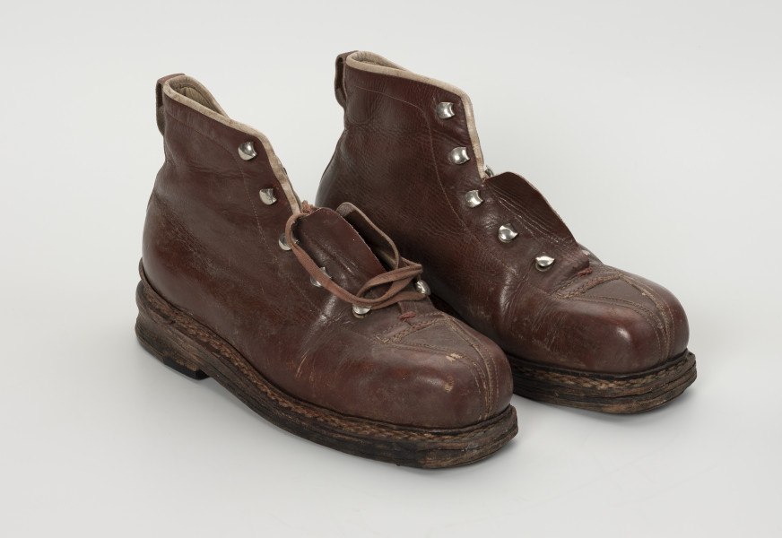 para trzewików - Ujęcie ze skosu z lewej; Para damskich, brązowych, sznurowanych butów. Otwory na sznurówki zabezpieczone metalowymi obwódkami. Cholewka wykonana ze skóry licowej bydlęcej. To naturalna skóra, której zewnętrzna warstwa została poddana obróbce mechanicznej powodującej jej gładką fakturę. Między płaty skóry wszyte skórzane wzmocnienia na noski. Podeszwy złożone z kilku warstw kruponu (najgrubszej części surowej skóry bydlęcej). Podeszwa zszywana. W wyniku zużycia skóry dosztukowane w latach późniejszych nowe warstwy kruponu z przodu podeszwy oraz fleki wykonane z gumy obuwniczej. Buty noszą ślady dużej eksploatacji, uszkodzenia skóry przy górnym zakończeniu buta oraz po całości widoczna popękana skóra.