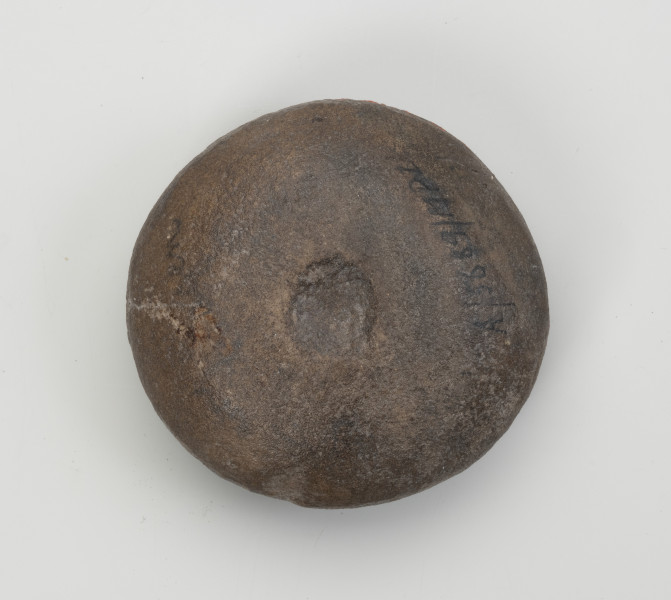 kamień kanelowany (kamień żłobkowany) - Ujęcie z dołu; Narzędzie kamienne z szarobrązowego otoczaka, widoczne koliste wgłębienie. Powierzchnia dość gładka, ślady obróbki widoczne na wgłębieniu.