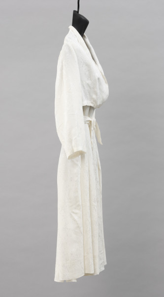 suknia ślubna - Ujęcie z prawej strony; Prosta suknia ślubna w kolorze białym, na szerokich ramiączkach, o długości do kolan, w pasie zwężona, dekolt głęboki. Spódnica z zakładkami rozkloszowana. Uszyta z jedwabnej żorżety, delikatnej tkaniny z nieregularnie marszczoną powierzchnią. Na całej powierzchni materiału wytłoczony wzór roślinny w postaci gałązek.