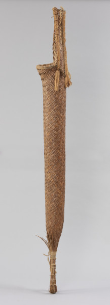 prasa do manioku - Ujęcie z boku; Długa, cylindryczna, pleciona prasa w formie wrzecionowatego wora. U szczytu posiada uchwyt,  od spodu jest związana. Wypleciona techniką taśmowo-krzyżową, dodatkowo wzmocniona: łykiem, sznurkiem z włókna roślinnego, w górnej części – wokół zewnętrznej krawędzi uchwytu i otworu – żyłką oraz w dolnej części – wokół pętli i powyżej niej – mocowaniem z naturalnego sznurka. Z zewnątrz ma naturalny kolor trzciny.