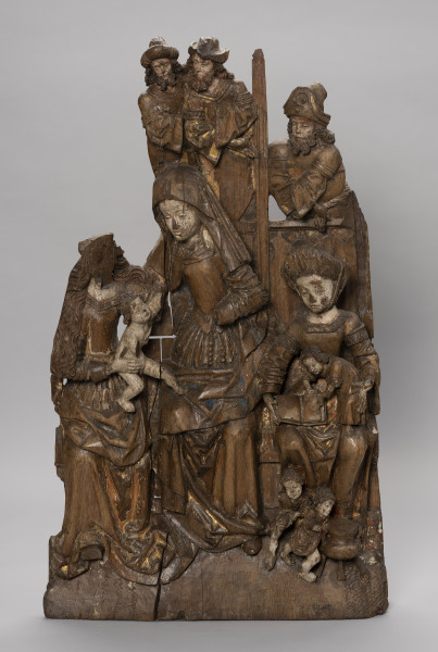 płaskorzeźba ołtarzowa; Rodzina Marii - Ujęcie z przodu; Wysoka płaskorzeźbiona płyta z przedstawieniem wielofigurowym, polichromowana. Ukazuje grupę strojnie ubranych kobiet siedzących na ławie i piastujących małe dzieci oraz mężczyzn widocznych powyżej wysokiego zaplecka ławy. W scenie dominuje przedstawienie św. Anny o nieco zwiększonych proporcjach. Święta w chuście na głowie i z różańcem przypiętym do paska wraz z Marią o długich falujących włosach podtrzymują nagiego Jezusa balansującego pomiędzy ich kolanami. Po prawej siedzi Maria Kleofasowa w wysokim marszczonym czepcu z czwórką synów ubranych w tuniki, z których jeden, stojący na kolanach, bawi się otwartą księgą, pozostali biegają wokół kociołka u stóp matki. Brodaci mężczyźni usytuowani za ławą, powyżej kobiet, widoczni w półpostaci, noszą tuniki na koszulach o szerokich rękawach i kunsztownie kształtowane kapelusze. Marszczone suknie z wąskim gorsetem i dekoltami w karo czy obszycia rękawów dekoracyjnymi taśmami, szerokie rękawy koszul to elementy strojów zgodne z niemiecką modą mieszczańską z początku XVI wieku. Modne stroje, łagodne wizerunki okrągłych twarzy a także poruszone pozy i gesty postaci nadają przedstawieniu charakter rodzajowy. Płaskorzeźba wybrana z łączonych bloków drewna, w dolnej części drążona. Płyta częściowo uszkodzona i popękana; brakuje postaci Marii Salome z dziećmi i mężem, męskich wizerunków powyżej Marii a także jej twarzy oraz czwartego dzieciątka u stóp Marii Salome. Polichromia zachowana szczątkowo.