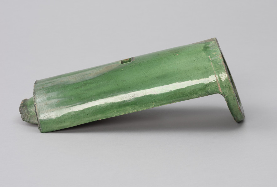 gąsior początkowy ozdobny - Ujęcie z boku; Półcylindryczna dachówka typu gąsior z zewnątrz szkliwiona zieloną glazurą. Zasklepiona z prawej okrągłą plakietką. Kwadratowy otwór wykonano w górnym grzbiecie dachówki. Na dachówce widoczne przetarcia, rysy, ubytki, spękania oraz odbarwienia - największe w górnej części plakietki i na grzbiecie – u dołu, przy krawędziach.