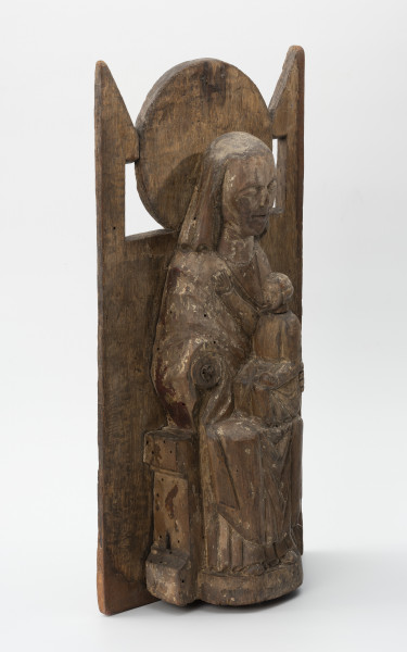 Tronująca Madonna z Dzieciątkiem - Ujęcie ze skosu z lewej; Niewielka figura tronującej Marii z dzieciątkiem o zwartej bryle, przytwierdzona do wysokiego zaplecka o geometrycznym zwieńczeniu, polichromowana. Rzeźba w typie Sedes Sapientiae ukazuje frontalnie siedzącą na tronie Marię w sztywnej, wyprostowanej pozie z dzieckiem w wieku chłopięcym usytuowanym bokiem, w pozycji siedzącej, ponad jej lewym kolanem. Długa suknia i płaszcz Marii oraz tunika Jezusa gładko przylegają do ciał postaci tworząc nieznaczne, schematyczne draperie w partii nóg. Między goleniami Marii tkanina układa się w charakterystyczne trójkątne fałdy o ostrych krawędziach. Hieratyczne pozy postaci przełamuje gest prawa ręka chłopca wyciągnięta wysoko i ułożona na piersi matki. Głowa Marii osadzona na sztywnej, szerokiej szyi okryta chustą ułożoną w tylnej partii na kształt welonu sięgającego do ramion. Twarz pociągła, owalna, zastygła w pozbawionym emocji wyrazie. Symetryczna fryzura chłopca z falujących włosów sięga poniżej uszu. Zwieńczenie gładko opracowanego zaplecka tworzy kolisty nimb za głową Marii flankowany trójkątnymi sterczynami oparcia tronu. Rzeźba opracowana jako przyścienna. Brak prawej dłoni Marii. Powierzchniowe ubytki drewna i otwory po drewnojadach. Polichromia zachowana szczątkowo.