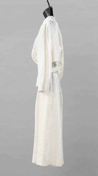 suknia ślubna - Ujęcie z lewej strony; Prosta suknia ślubna w kolorze białym, na szerokich ramiączkach, o długości do kolan, w pasie zwężona. Spódnica z zakładkami rozkloszowana. Uszyta z jedwabnej żorżety, delikatnej tkaniny z nieregularnie marszczoną powierzchnią. Na całej powierzchni materiału wytłoczony wzór roślinny w postaci gałązek.