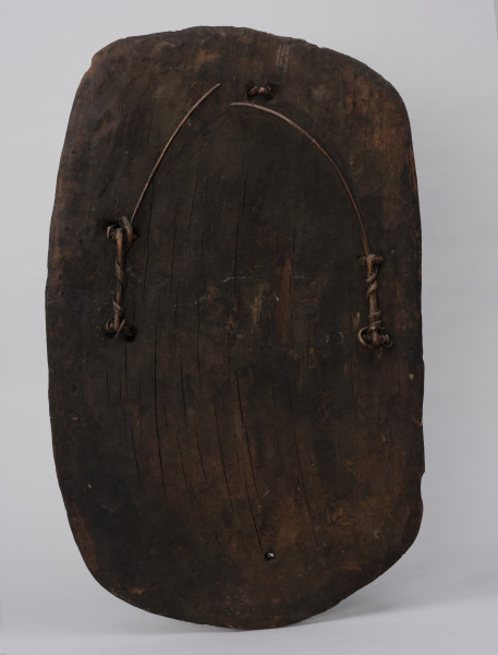 tarcza wojenna - Ujęcie z tyłu; Duża, drewniana, owalna tarcza prawdopodobnie przyciemniana sadzą. Na rewersie widać cztery pętle, przez które przechodzi zerwany imacz wykonany z takiego samego sznura. Przez pętlę znajdującą się u góry tarczy prawdopodobnie również przechodził sznurowy imacz, jednak z powodu jego uszkodzenia (zerwania) obecnie pozostaje pusta. Na powierzchni tarczy widoczne są większe i mniejsze spękania drewna, ślady ciosania wykonane kamiennym lub metalowym narzędziem oraz niewielkie wgłębienia po uszkodzeniach bojowych (strzałach).