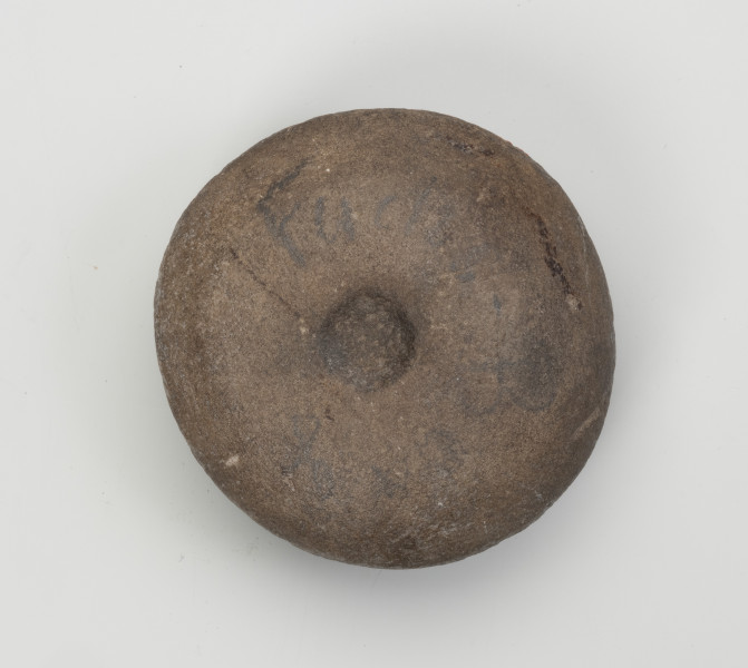 kamień kanelowany (kamień żłobkowany) - Ujęcie z góry; Narzędzie kamienne z szarobrązowego otoczaka, widoczne koliste wgłębienie. Powierzchnia dość gładka, ślady obróbki widoczne na całej powierzchni.