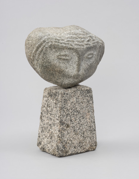 rzeźba; głowa - Ujęcie ze skosu z lewej; Szeroka, spłaszczona, bliska owoidalnej głowa dołem lekko wydłużona, osadzona na kostce w kształcie ściętego ostrosłupa. Rysy twarzy: łuki brwiowe, nos, konturowo zaznaczone oczy, drobne usta – ciemniejsze, wydobyte przez delikatny, negatywowy szlif policzków i obszarów wokół oczu. Ryt ornamentalny włosów widoczny tylko na froncie skały, bezpośrednio wokół twarzy.