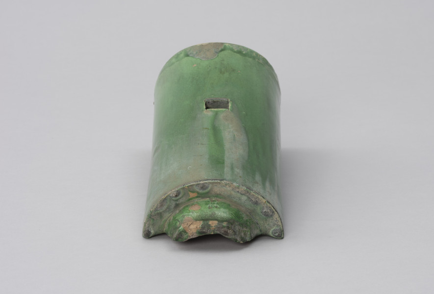 gąsior początkowy ozdobny - Ujęcie z dołu; Półcylindryczna dachówka typu gąsior z zewnątrz szkliwiona zieloną glazurą. Kwadratowy otwór wykonano w górnym grzbiecie dachówki. Na dachówce widoczne przetarcia, rysy, ubytki, spękania oraz odbarwienia - największe w górnej części plakietki i na grzbiecie – u dołu, przy krawędziach.