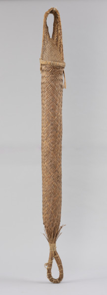 prasa do manioku - Ujęcie z tyłu; Długa, cylindryczna, pleciona prasa w formie wrzecionowatego wora. U szczytu posiada uchwyt w formie ucha, od spodu jest związana i zakończona rodzajem pętli. Wypleciona techniką taśmowo-krzyżową, dodatkowo wzmocniona: łykiem, sznurkiem z włókna roślinnego, w górnej części – wokół zewnętrznej krawędzi uchwytu i otworu – żyłką oraz w dolnej części – wokół pętli i powyżej niej – mocowaniem z naturalnego sznurka. Z zewnątrz ma naturalny kolor trzciny.