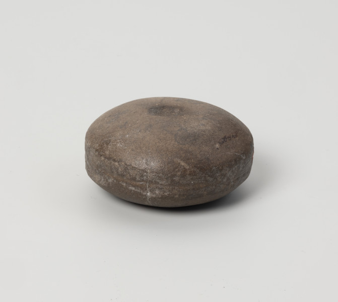 kamień kanelowany (kamień żłobkowany) - Ujęcie z przodu; Narzędzie kamienne z szarobrązowego otoczaka, w kształcie spłaszczonej kuli, na której obu biegunach widnieją koliste wgłębienia, na tym ujęciu widoczny tylko górny, natomiast po obwodzie biegnie delikatnie zarysowany szeroki żłobek. Powierzchnia dość gładka, ślady obróbki widoczne na wgłębieniach biegunów i ściankach bocznych.