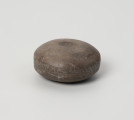 kamień kanelowany (kamień żłobkowany) - Ujęcie z przodu; Narzędzie kamienne z szarobrązowego otoczaka, w kształcie spłaszczonej kuli, na której obu biegunach widnieją koliste wgłębienia, na tym ujęciu widoczny tylko górny, natomiast po obwodzie biegnie delikatnie zarysowany szeroki żłobek. Powierzchnia dość gładka, ślady obróbki widoczne na wgłębieniach biegunów i ściankach bocznych.