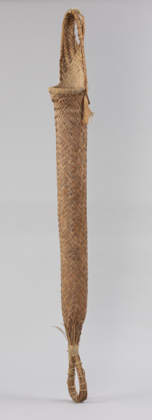 prasa do manioku - Ujęcie ze skosu z prawej; Długa, cylindryczna, pleciona prasa w formie wrzecionowatego wora. U szczytu posiada uchwyt w formie ucha, od spodu jest związana i zakończona rodzajem pętli. Wypleciona techniką taśmowo-krzyżową, dodatkowo wzmocniona: łykiem, sznurkiem z włókna roślinnego, w górnej części – wokół zewnętrznej krawędzi uchwytu i otworu – żyłką oraz w dolnej części – wokół pętli i powyżej niej – mocowaniem z naturalnego sznurka. Z zewnątrz ma naturalny kolor trzciny.