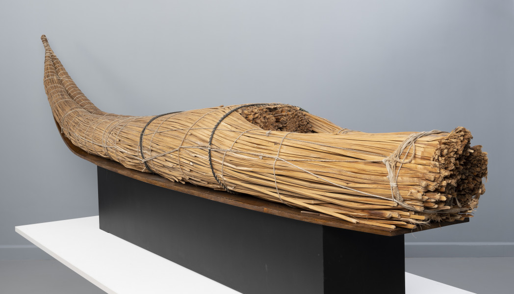 caballito de totora (łódź trzcinowa) - Ujęcie ze skosu z prawej; Ponad czterometrowa łódź trzcinowa o naturalnym jasnobrązowym kolorze z wysoko zadartym ku górze długim, zwężającym się dziobem oraz grubą, płasko ściętą rufą. Kształtem przypomina gruby cios słonia. Została zbudowana z dwóch wiązek trzciny mocno skrępowanych przez poprzeczne wiązania. W ten sposób nadano łodzi pożądany, żeglowny kształt. Wśród wiązek trzcinowych tworzących rufę uformowano trójkątne zagłębienie służące do przechowywania narzędzi rybackich (sieci, lin, pułapek) i złowionych zdobyczy.