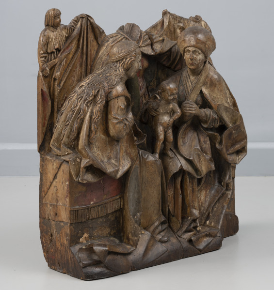 grupa rzeźbiarska - Ujęcie ze skosu z lewej; Grupa rzeźbiarska o zwartej, symetrycznej kompozycji, wykonana z jednego bloku drewna i polichromowana. Przedstawia trzy siedzące kobiety zwrócone ku dzieciątku stojącemu w centrum na ich kolanach. Analogicznie upozowane w układzie antytetycznym sylwetki długowłosej Marii po lewej stronie i Anny w czepcu po prawej okrywają obszerne płaszcze, układające się wokół figur dużymi płaszczyznami i opadające na podstawę sceny. Na ich kolanach stoi nagie dzieciątko o gładko opracowanej główce. Za nim, widoczna pomiędzy kobietami, siedzi frontalnie Emerancja o nieco powiększonych proporcjach, w prawej dłoni trzymając fragment pnia. Tło stanowi kotara unoszona przez dwa anioły widoczne w półpostaci, ubrane w szerokie tuniki. Tron okrywa tkanina obszyta frędzlami. Cechy anatomiczne postaci i modelunek ciężkich obszernych tkanin oddane są naturalistycznie. Rzeźba posiada znaczące ubytki – brakuje głów Emerancji i prawego anioła, dłoni Marii i prawej rączki dzieciątka oraz fragmentu podstawy z prawej strony. Na powierzchni rzeźby widoczne otwory po owadach. Polichromia zachowana częściowo.