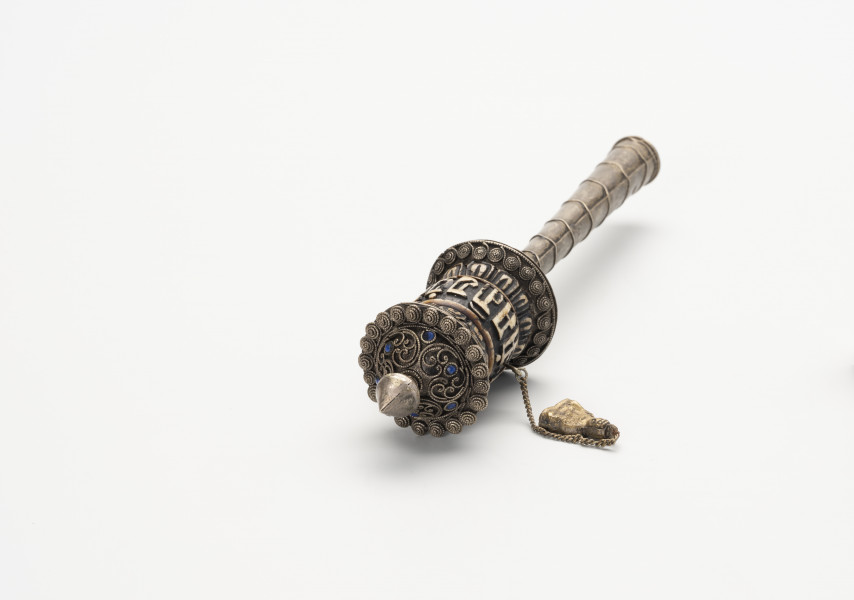 młynek modlitewny wraz z tekstem modlitewnym - Ujęcie z góry; Klepsydrowy, ręczny młynek modlitewny z uchwytem w formie tulejki w kształcie ściętego, wydłużonego stożka składający się z się z pięciu elementów. Mosiężny pąk lotosu u szczytu, kościano-mosiężna, klepsydrowa część z łańcuszkiem i inskrypcją oraz mosiężna podkładka tworzą bęben młynka. Poniżej znajdują się trzy kolejne elementy: niewielka, kościana podkładka, mosiężny uchwyt, z którego wystaje ostry, stalowy szpikulec. Młynek ozdobiono sześcioma, kamieniami pokrytymi niebieską emalią oraz motywami ślimacznicy, roślinnym, stylizowanych przecinków, zaokrąglonych linii i okręgów, zlokalizowanymi na mosiężnej powłoce bębna. Wymienione wzory wykonano z niklowanej blaszki mosiężnej, pociętej na drobne paseczki, przypominają zdobienie granulacją. Na kościanej części bębna wyrzeźbiono dwa razy słowa mantry: 