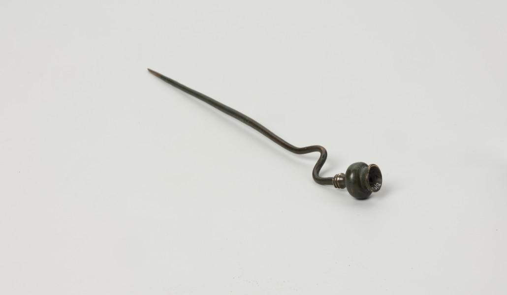 szpila z brązu ze Świchowa - Ujęcie z przodu; Szpila z brązu z wygiętą esowato  łabędzią szyjką. Zwieńczenie stanowi główka w formie miniaturowej wazy, której podstawę stanowią 3 dookolne zgrubienia w formie pierścieni. Powierzchnia zabytku jest w bardzo dobrym stanie, pokrywa ją ciemnozielona patyna.