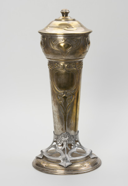 puchar klubu sportowego S.C. Blücher Stettin - ujęcie z tyłu; Metalowe naczynie z pokrywą i podstawą, wydłużone w pionie. Podstawa złożona z okrągłej, lekko wysklepionej stopy i wspartej na niej ażurowej, wykonanej z cyny części wspierającej czarę. Dolna część czary wyższa, smukła, cylindryczna rozszerza się ku górze, górna wybrzuszona część jest wydzielona od dołu i od góry pierścieniami. Pokrywa okrągła, wysklepiona, z uchwytem w formie gałki ozdobionej rozetką, na wierzchołku. Powierzchnia pucharu zdobiona wypukłym ornamentem secesyjnym. Na stopie profilowanie wokół otoku, a w górnej części potrójna linia dekoracji z czterema falistymi załamaniami. Analogiczny ornament zdobi pokrywę. Część ażurowa ukształtowana z asymetrycznych zwojów miękko wyciętej taśmy, przechodzącej w cztery nóżki mocowane na śruby do stopy i tyle samo wsporników korpusu, z których dwa przykryte są ułożonymi w bukiet trzema wielopłatkowymi kwiatkami. Ze środkowego kwiatka wyprowadzona jest symetryczna dekoracja dolnej części czary, złożona z ornamentu roślinno-geometrycznego: pionowy trzon rozdwaja się sercowo w wić zakończoną kwiatkami, wyżej powtórnie otacza pole o kształcie medalionu, a następnie wygina się i prowadzi poziomo wzdłuż poszerzenia czary wicią z kwiatami i owocami. W górnej części korpusu cztery symetryczne motywy ułożone w trójkątną formę z miękko wygiętych taśm.