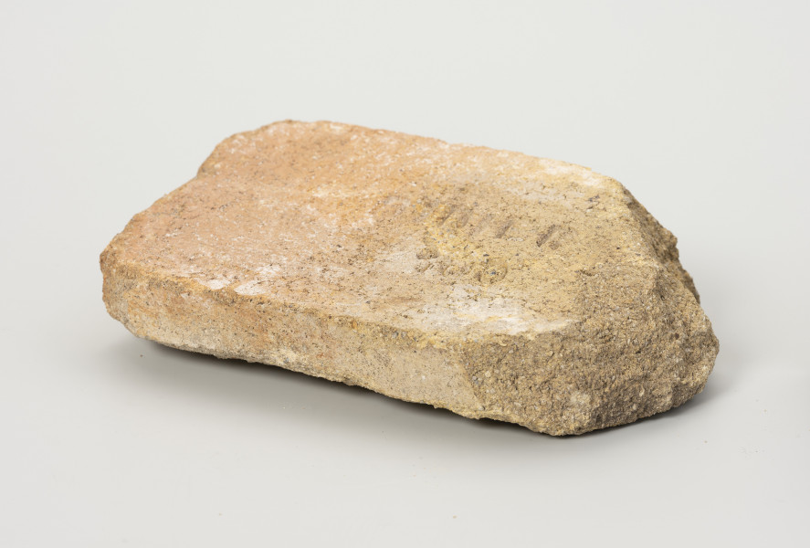 cegła szamotowa - Ujęcie przodu ze skosu z prawej; Prostopadłościenna cegła z jasnej glinki szamotowej. Prawa strona obtłuczona. Cegła niejednolita, ziarnista.