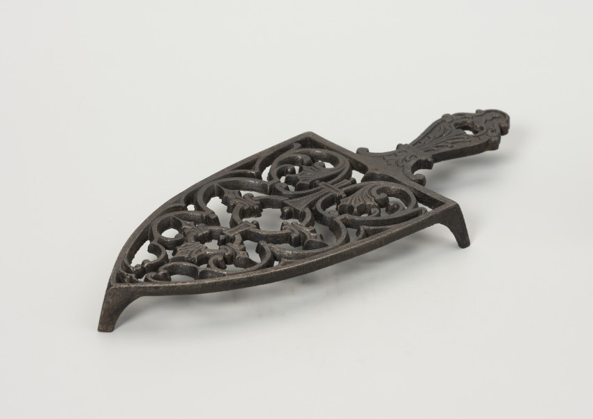podstawka pod żelazko - Ujęcie przodu ze skosu z prawej; Podstawka pod żelazko wykonana z żeliwa (odlew). Kształt zbliżony do trójkąta równoramiennego o lekko zaokrąglonych ramionach i prostej podstawie. Wnętrze podstawki ażurowe z wzorem roślinno-geometrycznym skomponowanym wzdłuż osi symetrii. Uchwyt płaski, profilowany z dekoracja roślinną i niewielkim otworem na końcu. Podstawka wsparta na zwężających się ku dołowi, niskich nóżkach.