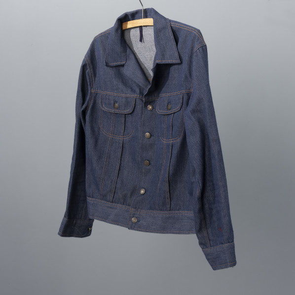 kurtka męska firmy ODRA - Ujęcie przodu ze skosu z prawej; Krótka, granatowa kurtka męska z materiału dżinsowego. Zapinana na guziki, z dwoma kieszeniami na klatce piersiowej posiadającymi klapki zapinane na guziki. Na całej kurtce widoczne szycie pomarańczową nitką. Każdy guzik posiada wypukłą głowę gryfa zwróconą w lewa stronę oraz na otoku napis ODRA SZCZECIN.