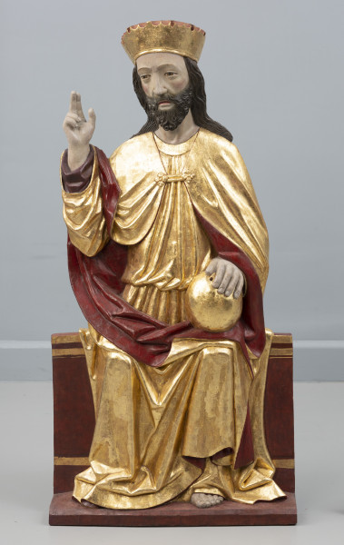 dwufigurowa grupa płaskorzeźb ołtarzowych - Ujęcie z przodu; Płaskorzeźbiona figura, na szerokiej ławie tronuje Chrystus. Postać ubrana jest w długą tunikę przewiązaną w pasie, płaszcz spięty klamrą powyżej piersi. Miękka, lejąca się ku podstawie rzeźby złocista tkanina,  szata połyskuje efektownie marszczeniami o łagodnych grzbietach. Na głowie wysoka złocona korona o gładkiej obręczy, zwieńczona zygzakiem. Chrystus swoją prawą rękę podnosi w geście błogosławieństwa, lewą opiera na złotej kuli trzymanej na kolanie. Jego pociągłą twarz o wyraźnie zaznaczonych kościach policzkowych pokrywa ciemny zarost. Długie włosy opadają na ramiona. Widoczne pęknięcie drewna na twarzy Chrystusa wzdłuż jego prawej skroni, poprzez policzek w kierunku ust. Fleurony wieńczące obręcze koron nie zachowały się. Złocenia płaskorzeźby częściowo rekonstruowane.