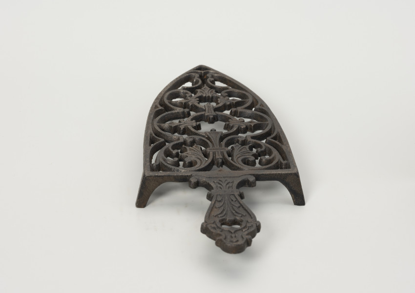 podstawka pod żelazko - Ujęcie z tyłu; Podstawka pod żelazko wykonana z żeliwa (odlew). Kształt zbliżony do trójkąta równoramiennego o lekko zaokrąglonych ramionach i prostej podstawie. Wnętrze podstawki ażurowe z wzorem roślinno-geometrycznym skomponowanym wzdłuż osi symetrii. Uchwyt płaski, profilowany z dekoracja roślinną i niewielkim otworem na końcu. Podstawka wsparta na trzech zwężających się ku dołowi, niskich nóżkach.