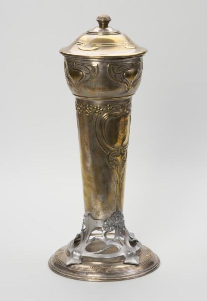 puchar klubu sportowego S.C. Blücher Stettin - ujęcie przodu ze skosu z lewej; Metalowe naczynie z pokrywą i podstawą, wydłużone w pionie. Podstawa złożona z okrągłej, lekko wysklepionej stopy i wspartej na niej ażurowej, wykonanej z cyny części wspierającej czarę. Dolna część czary wyższa, smukła, cylindryczna rozszerza się ku górze, górna wybrzuszona część jest wydzielona od dołu i od góry pierścieniami. Pokrywa okrągła, wysklepiona, z uchwytem w formie gałki ozdobionej rozetką, na wierzchołku. Powierzchnia pucharu zdobiona wypukłym ornamentem secesyjnym. Na stopie profilowanie wokół otoku, a w górnej części potrójna linia dekoracji z czterema falistymi załamaniami. Analogiczny ornament zdobi pokrywę. Część ażurowa ukształtowana z asymetrycznych zwojów miękko wyciętej taśmy, przechodzącej w cztery nóżki mocowane na śruby do stopy i tyle samo wsporników korpusu, z których dwa przykryte są ułożonymi w bukiet trzema wielopłatkowymi kwiatkami. Ze środkowego kwiatka wyprowadzona jest symetryczna dekoracja dolnej części czary, złożona z ornamentu roślinno-geometrycznego: pionowy trzon rozdwaja się sercowo w wić zakończoną kwiatkami, wyżej powtórnie otacza pole o kształcie medalionu, a następnie wygina się i prowadzi poziomo wzdłuż poszerzenia czary wicią z kwiatami i owocami. W centrum medalionu grawerowana tarcza z głową gryfa i napisem S.C. BLÜCHER STETTIN. W górnej części korpusu cztery symetryczne motywy ułożone w trójkątną formę z miękko wygiętych taśm; u dołu w obrębie pozbawionego dekoracji pola grawerowany napis: Ortsgruppe Stettin.