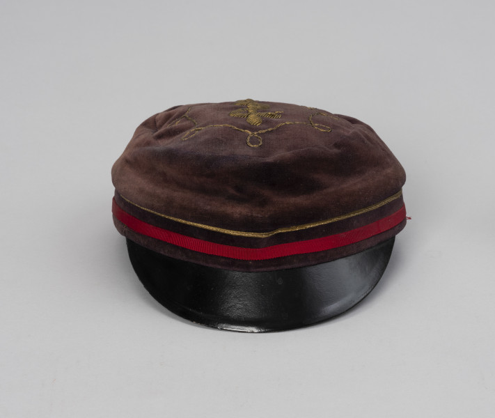 czapka studencka - Ujęcie z przodu; Czapka męska z brązowego aksamitu o okrągłym denku, z czarnym plastikowym daszkiem. Na denku, w przedniej części czapki wyszyty żółtą nicią wąż owinięty spiralnie na lasce.