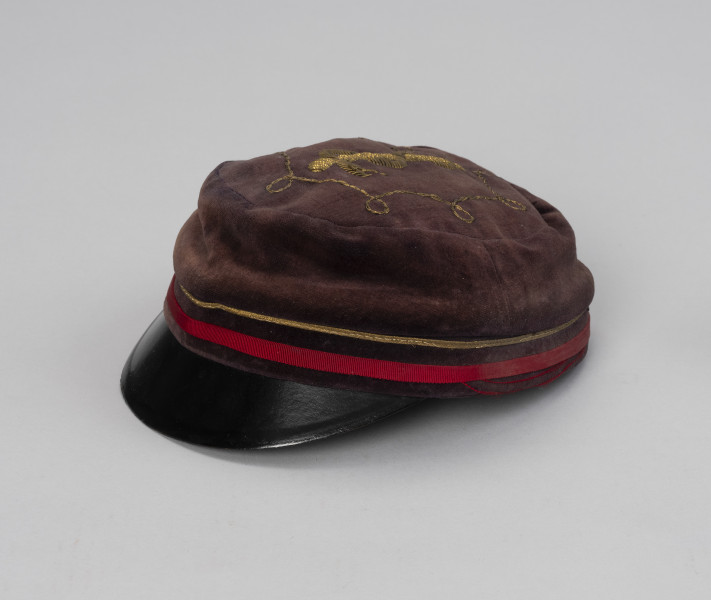 czapka studencka - Ujęcie przodu ze skosu z prawej; Czapka męska z brązowego aksamitu o okrągłym denku, z czarnym plastikowym daszkiem. Na denku, w przedniej części czapki wyszyty żółtą nicią wąż owinięty spiralnie na lasce.
