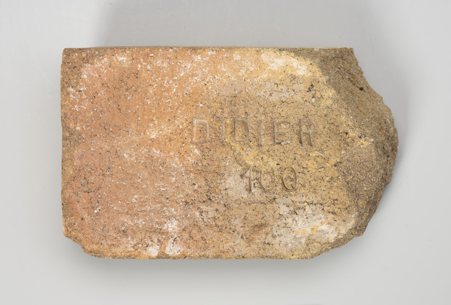 cegła szamotowa - Ujęcie z góry; Prostopadłościenna cegła z jasnej glinki szamotowej. Prawa strona obtłuczona. Cegła niejednolita, ziarnista. W centrum płaszczyzny ścianki poziomej tłoczony, wykonany metodą stempla, wklęsły napis DIDIER 100.