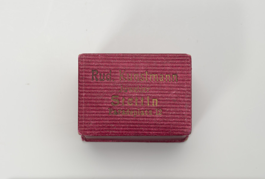 pudełko jubilerskie - Ujęcie z góry; Małe, prostopadłościenne pudełko na biżuterię, z twardej tektury w kolorze amarantowym, delikatnie prążkowane. Pudełeczko otwierane uchylnie. Na wieczku napis czcionką w kolorze złotym: Rud. Kunstmann Juwelier Stettin Paradeplatz 12.