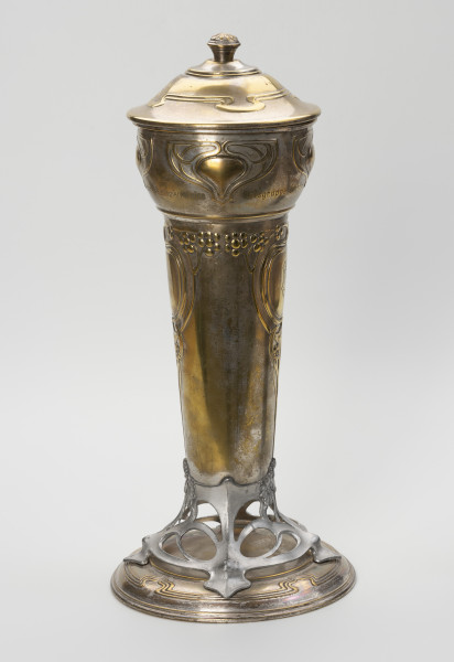 puchar klubu sportowego S.C. Blücher Stettin - ujęcie z boku; Metalowe naczynie z pokrywą i podstawą, wydłużone w pionie. Podstawa złożona z okrągłej, lekko wysklepionej stopy i wspartej na niej ażurowej, wykonanej z cyny części wspierającej czarę. Dolna część czary wyższa, smukła, cylindryczna rozszerza się ku górze, górna wybrzuszona część jest wydzielona od dołu i od góry pierścieniami. Pokrywa okrągła, wysklepiona, z uchwytem w formie gałki ozdobionej rozetką, na wierzchołku. Powierzchnia pucharu zdobiona wypukłym ornamentem secesyjnym. Na stopie profilowanie wokół otoku, a w górnej części potrójna linia dekoracji z czterema falistymi załamaniami. Analogiczny ornament zdobi pokrywę. Część ażurowa ukształtowana z asymetrycznych zwojów miękko wyciętej taśmy, przechodzącej w cztery nóżki mocowane na śruby do stopy i tyle samo wsporników korpusu, z których dwa przykryte są ułożonymi w bukiet trzema wielopłatkowymi kwiatkami. W górnej części korpusu cztery symetryczne motywy ułożone w trójkątną formę z miękko wygiętych taśm; u dołu w obrębie pozbawionego dekoracji pola grawerowany napis: Meisterschaft 1900-10 der I. Klasse des Verbandes Berliner Ballspielvereine oraz w dolnej linii Ortsgruppe Stettin.