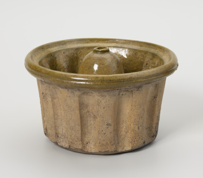 forma do wypieku babki - Ujęcie z przodu; Forma do wypieku babki o niewielkich rozmiarach i typowym dla tego rodzaju ciasta kształcie. Wysoki, prosty brzusiec regularnie karbowany i nieznacznie rozszerzający się ku górze przechodzi w wyraźnie wygięty na zewnątrz kołnierz o pogrubionej krawędzi. Centralna część dna wpuszczona do środka tworzy tak zwany komin (walec o kopulastym szczycie zakończonym małym otworkiem z zaznaczonym wylewem). Forma wykonana z gliny, wewnątrz jasnozielone szkliwo, zewnętrzna strona nie szkliwiona.