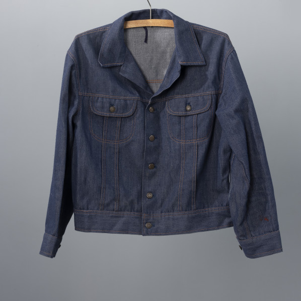 kurtka męska firmy ODRA - Ujęcie z przodu; Krótka, granatowa kurtka męska z materiału dżinsowego. Zapinana na guziki, z dwoma kieszeniami na klatce piersiowej posiadającymi klapki zapinane na guziki. Na całej kurtce widoczne szycie pomarańczową nitką. Każdy guzik posiada wypukłą głowę gryfa zwróconą w lewa stronę oraz na otoku napis ODRA SZCZECIN.