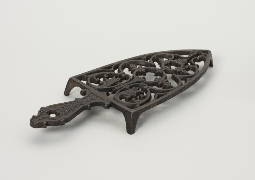 podstawka pod żelazko - Ujęcie tyłu ze skosu z prawej; Podstawka pod żelazko wykonana z żeliwa (odlew). Kształt zbliżony do trójkąta równoramiennego o lekko zaokrąglonych ramionach i prostej podstawie. Wnętrze podstawki ażurowe z wzorem roślinno-geometrycznym skomponowanym wzdłuż osi symetrii. Uchwyt płaski, profilowany z dekoracja roślinną i niewielkim otworem na końcu. Podstawka wsparta na trzech zwężających się ku dołowi, niskich nóżkach.