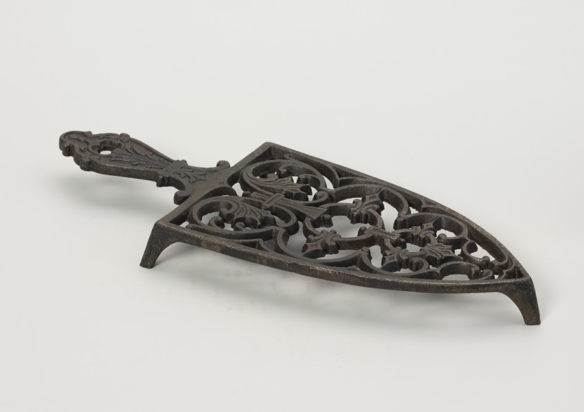 podstawka pod żelazko - Ujęcie przodu ze skosu z lewej; Podstawka pod żelazko wykonana z żeliwa (odlew). Kształt zbliżony do trójkąta równoramiennego o lekko zaokrąglonych ramionach i prostej podstawie. Wnętrze podstawki ażurowe z wzorem roślinno-geometrycznym skomponowanym wzdłuż osi symetrii. Uchwyt płaski, profilowany z dekoracja roślinną i niewielkim otworem na końcu. Podstawka wsparta na zwężających się ku dołowi, niskich nóżkach.
