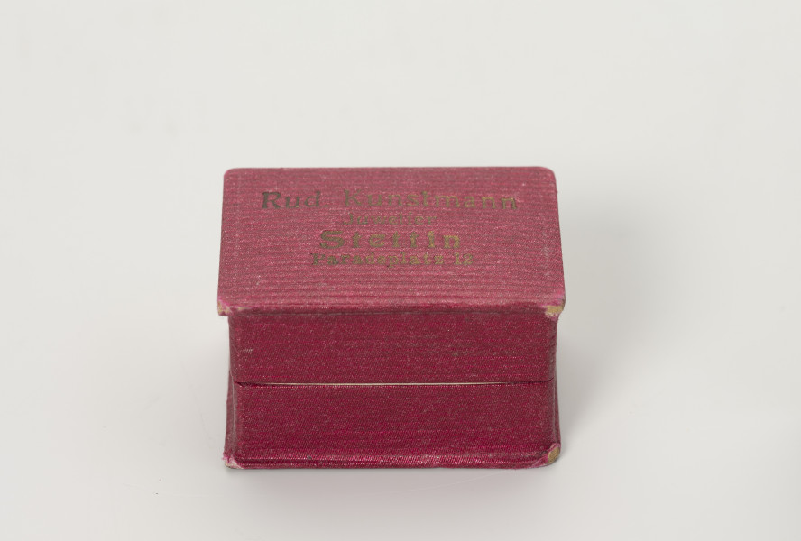 pudełko jubilerskie - Ujęcie z przodu; Małe, prostopadłościenne pudełko na biżuterię, z twardej tektury w kolorze amarantowym, delikatnie prążkowane. Pudełeczko otwierane uchylnie. Na wieczku napis czcionką w kolorze złotym: Rud. Kunstmann Juwelier Stettin Paradeplatz 12.