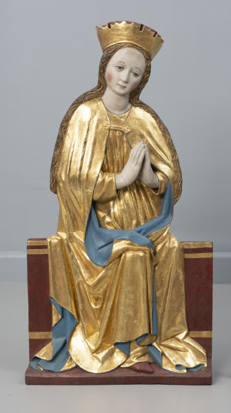 dwufigurowa grupa płaskorzeźb ołtarzowych - Ujęcie z przodu; Płaskorzeźbiona figura przedstawia na szerokiej ławie tronuje Maria. Postać ubrana jest w długą tunikę przewiązaną w pasie i płaszcz spięty klamrą powyżej piersi. Miękka, lejąca się ku podstawie rzeźby złocista tkanina, szata połyskuje efektownie marszczeniami o łagodnych grzbietach. Na głowie wysoka złocona korona o gładkiej obręczy, zwieńczona zygzakiem. Maria lekko przechyla głowę wlewo, dłonie trzymając w geście modlitewnym przy piersi. Jej młodzieńczą twarz o wysokim czole i łagodnych rysach otaczają pasma długich falujących, złocistych włosów.  Długie włosy opadają na ramiona. Tył rzeźby opracowany płasko. Fleurony wieńczące obręcze koron nie zachowały się. Złocenia płaskorzeźby częściowo rekonstruowane.