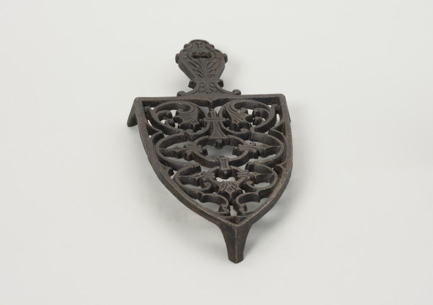 podstawka pod żelazko - Ujęcie z przodu; Podstawka pod żelazko wykonana z żeliwa (odlew). Kształt zbliżony do trójkąta równoramiennego o lekko zaokrąglonych ramionach i prostej podstawie. Wnętrze podstawki ażurowe z wzorem roślinno-geometrycznym skomponowanym wzdłuż osi symetrii. Uchwyt płaski, profilowany z dekoracja roślinną i niewielkim otworem na końcu. Podstawka wsparta na trzech zwężających się ku dołowi, niskich nóżkach.