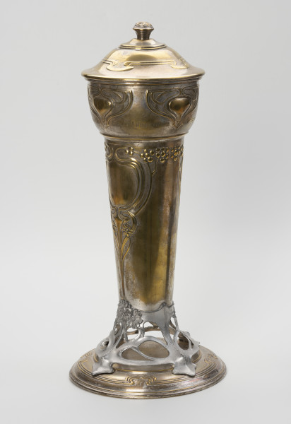 puchar klubu sportowego S.C. Blücher Stettin - ujęcie z boku; Metalowe naczynie z pokrywą i podstawą, wydłużone w pionie. Podstawa złożona z okrągłej, lekko wysklepionej stopy i wspartej na niej ażurowej, wykonanej z cyny części wspierającej czarę. Dolna część czary wyższa, smukła, cylindryczna rozszerza się ku górze, górna wybrzuszona część jest wydzielona od dołu i od góry pierścieniami. Pokrywa okrągła, wysklepiona, z uchwytem w formie gałki ozdobionej rozetką, na wierzchołku. Powierzchnia pucharu zdobiona wypukłym ornamentem secesyjnym. Na stopie profilowanie wokół otoku, a w górnej części potrójna linia dekoracji z czterema falistymi załamaniami. Analogiczny ornament zdobi pokrywę. Część ażurowa ukształtowana z asymetrycznych zwojów miękko wyciętej taśmy, przechodzącej w cztery nóżki mocowane na śruby do stopy i tyle samo wsporników korpusu, z których dwa przykryte są ułożonymi w bukiet trzema wielopłatkowymi kwiatkami. Ze środkowego kwiatka wyprowadzona jest symetryczna dekoracja dolnej części czary, złożona z ornamentu roślinno-geometrycznego: pionowy trzon rozdwaja się sercowo w wić zakończoną kwiatkami, wyżej powtórnie otacza pole o kształcie medalionu, a następnie wygina się i prowadzi poziomo wzdłuż poszerzenia czary wicią z kwiatami i owocami. W górnej części korpusu symetryczne motywy ułożone w trójkątną formę z miękko wygiętych taśm; u dołu w obrębie pozbawionego dekoracji pola grawerowany napis: Berliner Ballspielvereine.