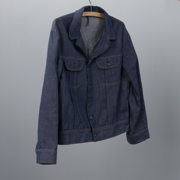 kurtka męska firmy ODRA - Ujęcie przodu ze skosu z lewej; Krótka, granatowa kurtka męska z materiału dżinsowego. Zapinana na guziki, z dwoma kieszeniami na klatce piersiowej posiadającymi klapki zapinane na guziki. Na całej kurtce widoczne szycie pomarańczową nitką. Każdy guzik posiada wypukłą głowę gryfa zwróconą w lewa stronę oraz na otoku napis ODRA SZCZECIN.